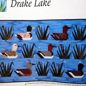 Puede incluir: Una colcha que representa una escena de lago con patos nadando en el agua y juncos verdes. La colcha se titula "Drake Lake" y está diseñada por Rebecca Manthey, cosida por Lucy Brown y acolchada por Maria Garm.