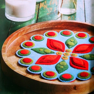 May include: A decorative felt coaster with a floral design in a wooden tray. The coaster features a light blue background with red, green, and orange felt appliqués. A lit candle in a glass jar is in the background.