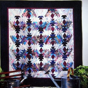 May include: A handmade quilt with a geometric pattern in shades of blue, purple, and burgundy. The quilt features a dark blue border and a complex design of diamond and triangle shapes. The quilt is displayed near a bench and watering can.