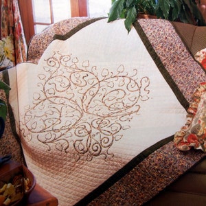 May include: A white quilt with a brown embroidered design of a tree with leaves. The quilt is on a sofa with a floral patterned fabric.