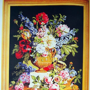 May include: A framed cross-stitch embroidery depicting a vase filled with a variety of colorful flowers, including roses, tulips, and lilies. The vase is decorated with gold accents and sits on a table with a brown wooden surface.