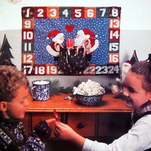 Può includere: Due bambini sono seduti a un tavolo con un calendario dell'avvento natalizio con due Babbo Natale che tengono bastoncini di zucchero e un cuore rosso. Il calendario ha i numeri da 1 a 24. I bambini stanno tenendo caramelle e popcorn.