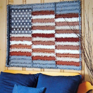 May include: A patriotic, handmade American flag quilt. The quilt features a blue field with white stars and alternating red, white, and patterned stripes. The edges are fringed, adding a rustic touch. The quilt is displayed on a wooden wall.