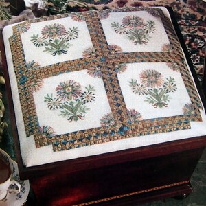 May include: A white fabric footstool with a wooden frame. The fabric is embroidered with four floral designs in shades of pink, green, and blue. The floral designs are surrounded by a geometric pattern in gold and blue.