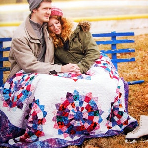 May include: A couple sits on a blue bench wrapped in a white quilt with a colorful patchwork design. The quilt has a large, circular pattern in the center, with smaller circles surrounding it. The couple is wearing winter clothing, and the woman is wearing a pink hat.