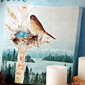 Puede incluir: Un tapiz de pared acolchado que representa un pájaro posado en una rama de árbol con un nido que contiene tres huevos azules. La obra de arte representa un paisaje sereno con un lago y un bosque, mostrando un tema inspirado en la naturaleza.