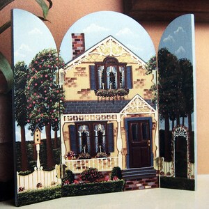 May include: A three-panel painted wooden screen depicting a yellow brick house with a blue door and white trim. The house has a porch with white railings and a flower box under the window. The screen is surrounded by a green lawn and trees.
