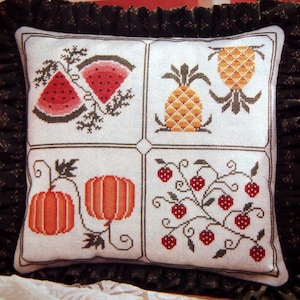 Puede incluir: Cojín cuadrado con fondo blanco con diseños de punto de cruz de rodajas de sandía, una piña, calabazas y fresas. El cojín tiene un borde negro con volantes.