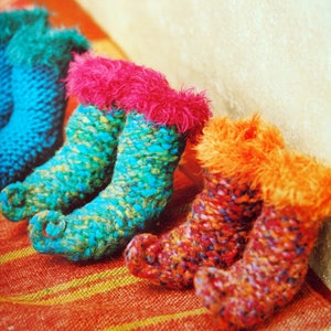May include: A selection of handmade elf boots in a row. The boots are knitted with textured yarn in shades of blue, teal, pink, and orange. Each boot has a fluffy trim around the top. The boots are arranged on a striped orange and yellow surface.