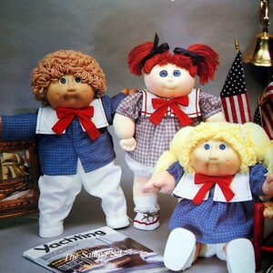 May include: Three Cabbage Patch Kids dolls. One doll has brown curly hair, blue and white outfit, and a red bow tie. Another doll has red pigtails, a red bow tie, and a blue and white plaid outfit. The third doll has blonde pigtails and a blue and white dress.