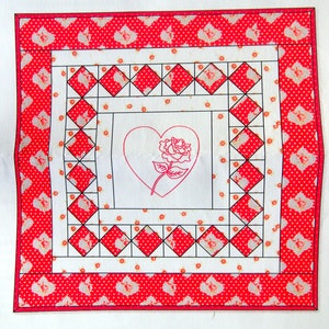 May include: A white fabric square with a red and white polka dot border. The center of the square features a black and white diamond pattern with a red heart and a rose in the center.