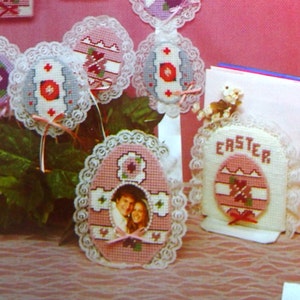 May include: A collection of plastic canvas Easter crafts featuring Victorian-inspired designs. The crafts include a tissue box cover, a photo frame, a card holder, and several ornaments. The ornaments feature a variety of Easter themes, including eggs, flowers, and bunnies. The crafts are all decorated with lace and ribbons.