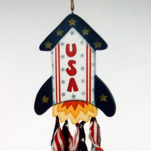 May include: A blue and white rocket-shaped wooden ornament with the word "USA" painted on it. The rocket has a red, white, and blue streamer tail.