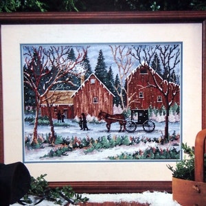 May include: A Dimensions cross stitch kit package titled Snowy Sunday by Al Koenig. The packaging features a framed cross stitch artwork of red barns, a horse-drawn carriage, and figures in a snowy landscape, with a black bonnet and wooden basket.