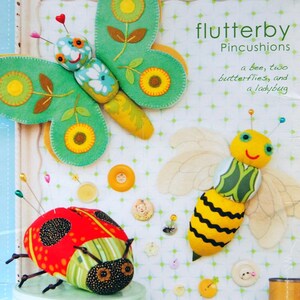 Peut inclure: Des coussins à épingles en feutre faits à la main en forme de papillon, d'abeille et de coccinelle. Le papillon est vert avec des détails jaunes et floraux. L'abeille est jaune et noire. La coccinelle est rouge avec des pois noirs. Le texte "flutterby Pincushions" est visible.