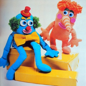 May include: Two handmade plush toys, one blue with a red hat and yellow bow tie, the other orange with a long orange nose and red cheeks. Both toys are sitting on yellow boxes.