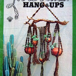 May include: A close-up of a macrame wall hanging with four plant hangers. The hangers are made of brown rope and have a natural wood branch at the top. The hangers are decorated with green plants and brown pots. The text "Macrame Hang Ups" is printed in black on a green background.