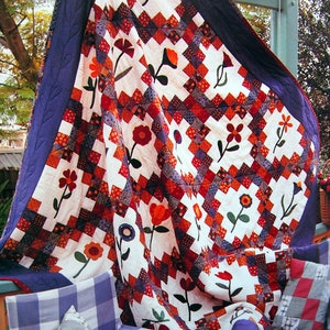 May include: A white quilt with a patchwork design featuring red, blue, and orange squares. The quilt is decorated with appliqued flowers in shades of red and orange. The quilt is draped over a chair with blue and white checkered cushions.