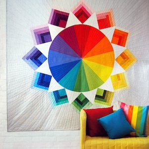 May include: A colourful quilt with a rainbow colour wheel design in the centre and a white background. The quilt is hanging on a white wall above a yellow armchair with colourful cushions.