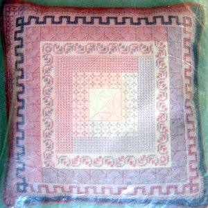 May include: A pink and purple embroidered pillow with a geometric pattern and a white border. The pillow is surrounded by a plastic bag.