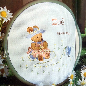 Peut inclure: Un cercle de broderie blanc avec un motif de point de croix représentant un ours en peluche portant une robe bleu et blanc et un chapeau bleu. L'ours tient une fleur et est entouré de petites fleurs blanches. Le texte "Zoe" et "23-6-95" est brodé en fil brun au-dessus de l'ours.