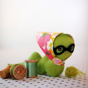 May include: A whimsical green plush creature wearing black glasses and a floral headscarf. Sewing notions, including spools of green thread, a thimble, and a ball of yarn, are arranged nearby. The background is a white surface with blue polka dots.