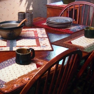 May include: A wooden table with a brown tablecloth and four placemats. Each placemat is a different pattern of fabric. There are two mugs, a bowl, and a set of plates on the table.