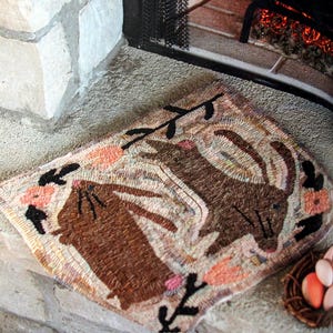 May include: A brown and cream rug with a design of two rabbits and floral accents. The rug is placed in front of a fireplace.