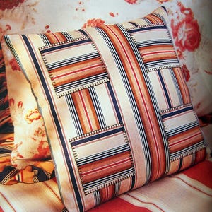 May include: A striped cushion with a patchwork design featuring multiple squares of striped fabric sewn together. The cushion is made of a white fabric with red, orange, and blue stripes.