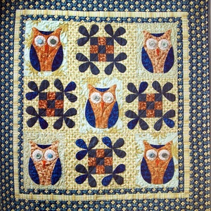 May include: A quilt with a patchwork design featuring owls with button eyes. The owls are surrounded by blue and brown floral shapes on a beige background. The quilt is bordered with a blue and white patterned fabric.