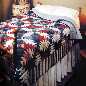 May include: A bed with a colourful quilt featuring a geometric pattern in red, white, blue, and green. The quilt is draped over a bed with a wooden frame and a white bed skirt. Decorative boxes are stacked on a nearby surface.