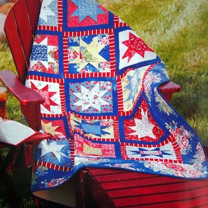 Op de afbeelding: Een rood, wit en blauw patchwork-deken met een sterpatroon is over een rode houten stoel gegooid. Het deken is gemaakt van verschillende stoffen met bloemen- en geometrische patronen.