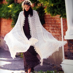 May include: A woman wearing a flowing, cream-colored lace shawl with intricate geometric patterns and a fringed edge. The shawl is draped over a dark top and a patterned skirt. The background includes a brick wall and greenery.