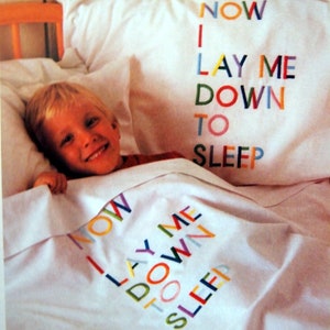 Op de afbeelding: Een jong kind ligt in bed met een wit laken en kussen. Het kussen heeft de woorden "NOW I LAY ME DOWN TO SLEEP" geborduurd in kleurrijke letters.