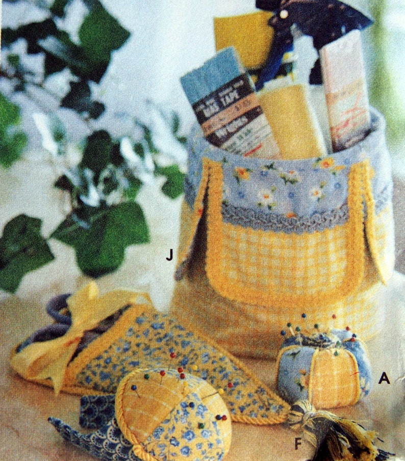 May include: A collection of sewing accessories, including a fabric basket, pin cushions, and a needle holder. The basket is blue and yellow, with a floral pattern and a gingham pocket. The pin cushions are round and teardrop-shaped, with pins inserted.