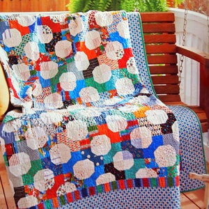 May include: A colorful patchwork quilt with a geometric pattern of octagons and squares. The quilt is made of various fabrics in shades of blue, red, white, and green. It is draped over a wooden porch swing.