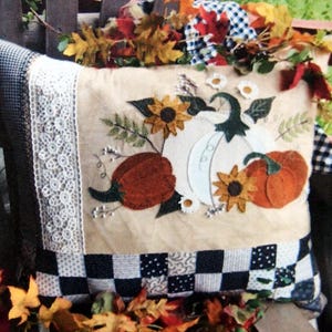 May include: A decorative pillow with a fall theme featuring a white pumpkin, two orange pumpkins, sunflowers, and leaves. The pillow has a black and white checkered fabric on the bottom and a white lace trim on the side.