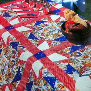 May include: A red and white checkered picnic blanket with a blue and white star pattern. The blanket is laid out on a grassy surface with a picnic basket, food, and drinks.