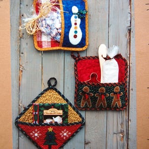 May include: Three quilted Christmas-themed pot holders. One features a snowman design, another has gingerbread men, and the third displays a box of cookies and a Christmas tree. Each has a loop for hanging.