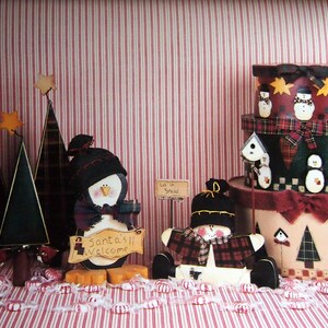 Puede incluir: Decoraciones navide&ntilde;as festivas que incluyen &aacute;rboles de madera con estrellas, un ping&uuml;ino con un letrero de "Santa's Welcome" y un mu&ntilde;eco de nieve. Cajas decorativas apiladas con dise&ntilde;os de mu&ntilde;ecos de nieve y casitas de p&aacute;jaros sobre un fondo a rayas rojas y blancas.