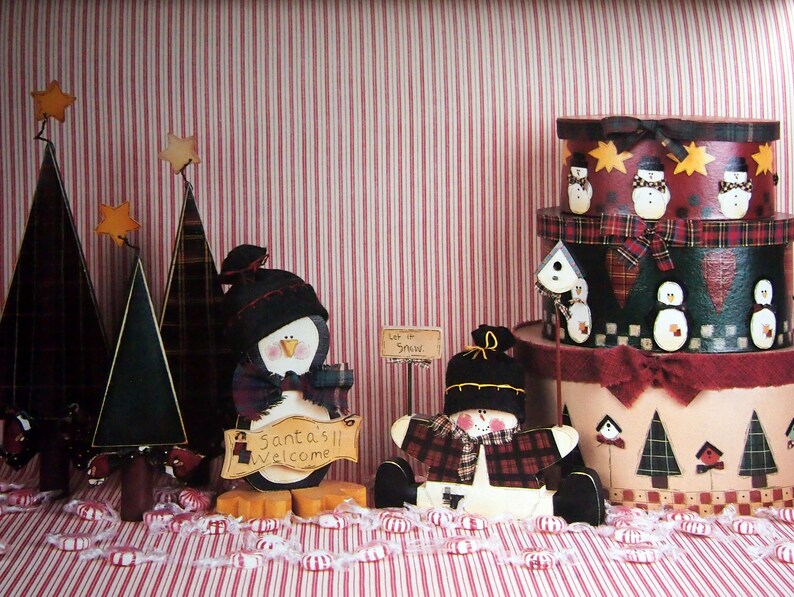 Puede incluir: Decoraciones navide&ntilde;as festivas que incluyen &aacute;rboles de madera con estrellas, un ping&uuml;ino con un letrero de "Santa's Welcome" y un mu&ntilde;eco de nieve. Cajas decorativas apiladas con dise&ntilde;os de mu&ntilde;ecos de nieve y casitas de p&aacute;jaros sobre un fondo a rayas rojas y blancas.