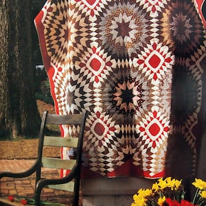 Puede incluir: Una colcha roja, blanca y marrón con un patrón de estrella estrellada cuelga de una pared. La colcha está hecha de tela de retazos y tiene un aspecto rústico y vintage. Una mecedora se encuentra frente a la colcha, y un ramo de flores amarillas y rojas se encuentra en el suelo.
