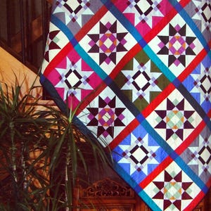 Peut inclure: Une courtepointe colorée avec un motif géométrique d'étoiles et de carrés dans des tons de rouge, bleu, vert, blanc et noir. La courtepointe est accrochée à un escalier en bois.