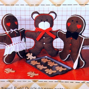 May include: A set of three plush toys: a gingerbread person wearing a white apron, a gingerbread person wearing a black bow tie, and a brown teddy bear wearing a red and white polka dot bow. The toys are sitting on a red surface with a baking sheet of gingerbread cookies.