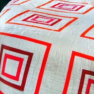 May include: White quilt with a geometric pattern of red and pink squares. The squares are outlined in red and pink, creating a layered effect.