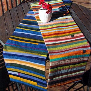 May include: Two colorful, striped table runners in shades of blue, green, orange, and red. A white vase with red flowers sits on one of the runners. The runners are placed on a dark wooden table.