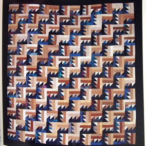 May include: A handmade quilt with a geometric pattern. The quilt features a repeating design of angular shapes in shades of brown, beige, blue, and black. The quilt is framed by a solid black border, creating a striking contrast.