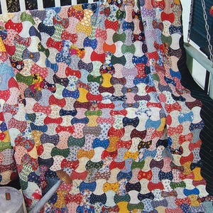 May include: A patchwork quilt featuring a repeating spool or bowtie pattern composed of numerous small fabric squares in a wide variety of colors, including red, blue, yellow, green, and white, with diverse floral, polka dot, and geometric prints.