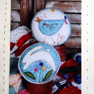 May include: Two felt pincushions, one shaped like a bird with a blue body and the other shaped like a rabbit with a white body. Both pincushions have a blue background with embroidered details. The pincushions are sitting on a red and white quilt.