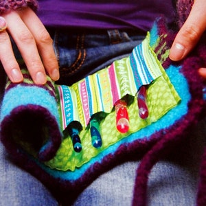 Può includere: Un astuccio in feltro fatto a mano con un interno in tessuto a righe colorate. L'astuccio ha un esterno blu, viola e verde con diverse perline di vetro colorate all'interno. L'astuccio è tenuto da una persona che indossa un maglione viola.